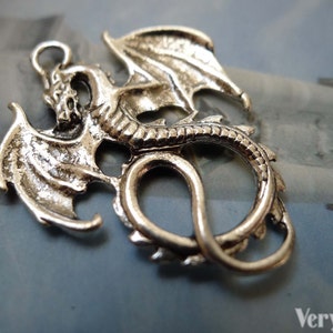 May include: Silver-toned metal dragon charm with detailed wings and a curled tail. The dragon is in a coiled position with its wings spread out.