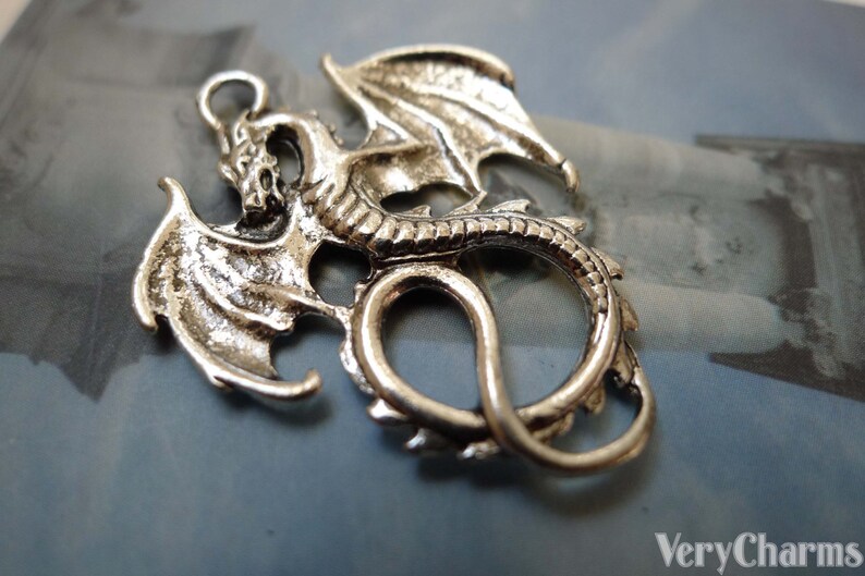 May include: Silver-toned metal dragon charm with detailed wings and a curled tail. The dragon is in a coiled position with its wings spread out.