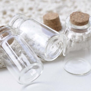 May include: Three small clear glass bottles with cork stoppers. The bottles are empty and sitting on a white crocheted surface.