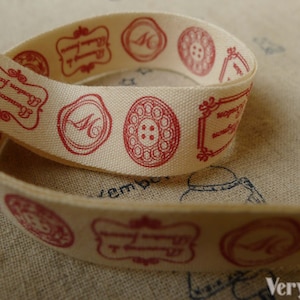 Puede incluir: Cinta de tela de color crema con diseños impresos en rojo. Los diseños incluyen un teléfono, una letra "M", una flor y una forma geométrica. La cinta está doblada por la mitad.