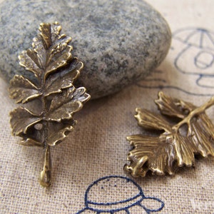 May include: Two bronze-colored leaf charms with intricate veining details. One charm is upright, while the other lies on its side. The charms are set against a neutral background with a gray stone.