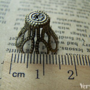 May include: A close-up of a small, ornate, gold-toned metal cap with a decorative design. The cap is about 1 cm in diameter and has a scalloped edge. It is sitting on a ruler with markings in centimeters.