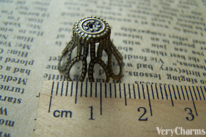 May include: A close-up of a small, ornate, gold-toned metal cap with a decorative design. The cap is about 1 cm in diameter and has a scalloped edge. It is sitting on a ruler with markings in centimeters.