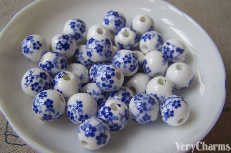Puede incluir: Una colecci&oacute;n de cuentas de cer&aacute;mica blanca con un patr&oacute;n floral azul. Las cuentas son redondas y tienen un agujero en el centro para enhebrar.