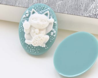 May include: Two oval-shaped, light blue resin cabochons. One features a white, raised, three-dimensional design of a Japanese Maneki-Neko, or lucky cat, with a raised paw. The other cabochon is plain.