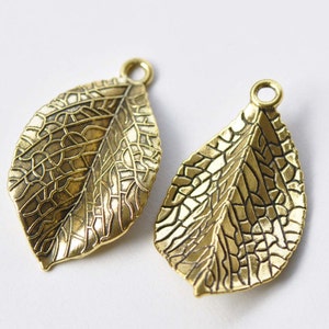 May include: Two gold-toned metal leaf charms with a detailed textured design. The leaves have a slightly pointed tip and a hole at the top for attaching to jewelry or crafts.