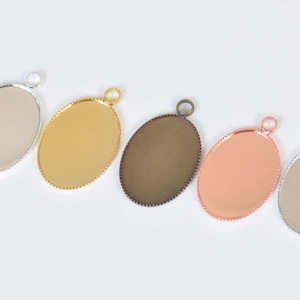 May include: Five oval-shaped metal pendant trays with a beaded edge. The trays are silver, gold, bronze, rose gold, and silver. Each tray has a small loop at the top for attaching a chain or cord.
