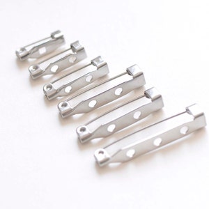 May include: A set of six silver-toned metal safety pin closures for crafts and jewelry making. The pins are arranged in order of size, with the smallest pin at the top and the largest pin at the bottom.