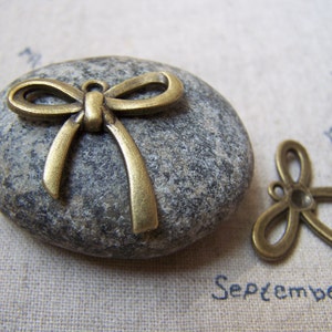 May include: A bronze-colored metal bow charm with a loop for hanging. The charm is sitting on a gray stone.