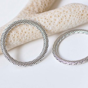 May include: Two silver rings with a textured, woven design. The rings are on a white background.