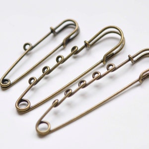 May include: Three antique brass safety pins arranged on a white surface. Each pin has a classic design with a rounded head and a series of decorative loops along the shaft. The pins are slightly angled, showcasing their vintage aesthetic.