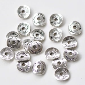 May include: A collection of silver-toned metal beads with a sunburst design. The beads are flat and have a hole in the centre for stringing.