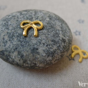 Könnte beinhalten: Zwei goldfarbene Metall-Schleifen-Anhänger. Die größere Schleife befindet sich auf einem grauen Stein. Die kleinere Schleife befindet sich auf einem weißen Stoffhintergrund.