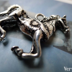 May include: A silver-colored horse pendant in a rearing pose. The detailed charm features a textured surface and a small loop for attaching to a necklace or bracelet. The horse is a symbol of freedom and power.