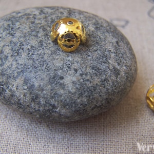 May include: Two gold-colored metal bead caps with a decorative openwork design. The caps are shown on a gray rock.