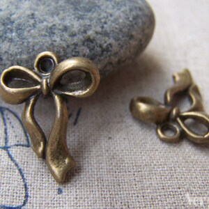 May include: Two antique gold-toned metal bow charms. The larger bow charm has a loop at the top for attaching to a necklace or bracelet. The smaller bow charm has a more intricate design with a loop at the bottom.