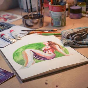 Peut inclure: Aquarelle d'une figure allong&eacute;e sur un bloc de papier blanc, sur un cercle vert. L'&oelig;uvre est sur une table en bois, entour&eacute;e de fournitures d'art comme des tubes de peinture, des pinceaux et un pot. Une petite impression de l'&oelig;uvre est &eacute;galement visible.
