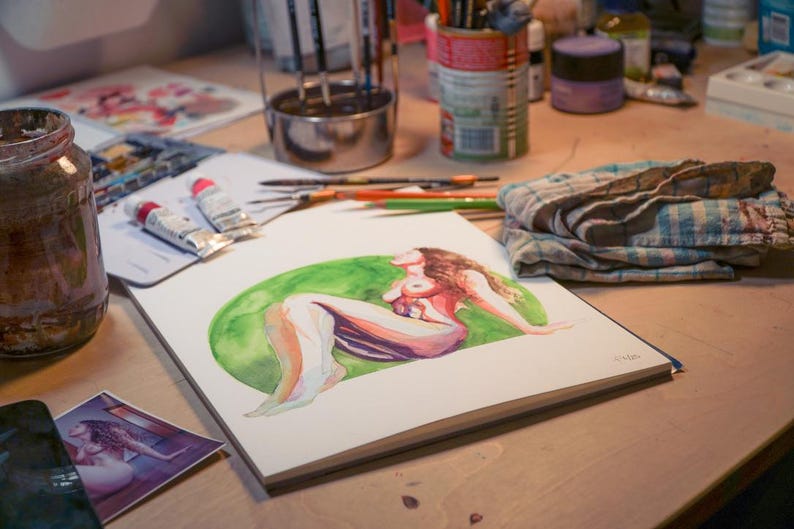 Peut inclure: Aquarelle d'une figure allong&eacute;e sur un bloc de papier blanc, sur un cercle vert. L'&oelig;uvre est sur une table en bois, entour&eacute;e de fournitures d'art comme des tubes de peinture, des pinceaux et un pot. Une petite impression de l'&oelig;uvre est &eacute;galement visible.