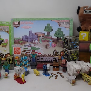 May include: A collection of Minecraft-themed toys and plushies. Includes building sets, character figurines, and plush animals like a brown cow and a green Creeper. A yellow question mark block and a Minecraft logo are also visible.