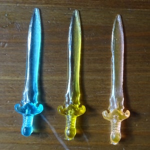 May include: Three plastic swords in different colors: blue, yellow, and pink. The swords are translucent and have a pointed tip.