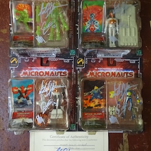 May include: Four sealed plastic packages containing action figures from the Micronauts toy line. Each package features a different character, including Acroyear, Space Glider, and two others. The packages are signed by the artist, Michael Horn, President and CEO of Palisades Toys.