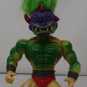 May include: Green and purple action figure with a green mohawk and a red loincloth. The figure has a fierce expression and is posed in a fighting stance.