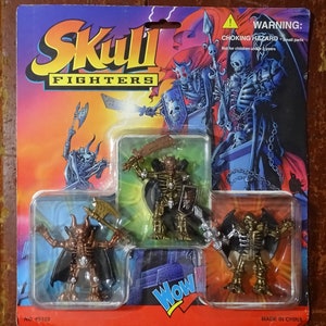 Puede incluir: Un juego de figuras de acción Skull Fighters en un blíster de plástico. El embalaje es azul, morado y rojo, con ilustraciones de guerreros esqueleto. El juego incluye tres figuras de esqueleto con armas y capas. La etiqueta de advertencia indica "PELIGRO DE ASFIXIA - Piezas pequeñas".
