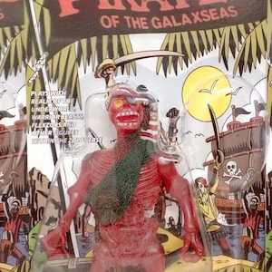 Könnte beinhalten: Eine rote und grüne Plastik-Actionfigur eines Skelettpiraten mit einem Totenkopf-Augenklappen. Die Figur heißt "Patch Plankwalker" und ist Teil der Spielzeuglinie "Pirates of the Galaxseas". Die Figur ist in einer Plastik-Blisterverpackung mit einem bunten Hintergrund verpackt.