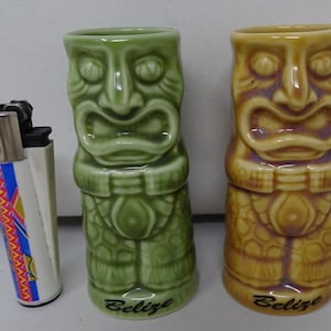 Pode incluir: Dois copos de shot tiki de cerâmica com esmalte verde e marrom. Os copos têm a forma de figuras tiki com rostos e têm a palavra "Belize" impressa na parte inferior.