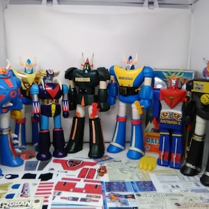 May include: A collection of vintage robot toys, including a blue and red robot with a red chest plate, a black and yellow robot with a yellow chest plate, a green robot with a black chest plate, a blue robot with a yellow chest plate, a red and white robot with a red chest plate, and a black robot with a yellow chest plate. The robots are all standing on a white surface with various packaging and accessories.