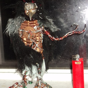 May include: A black and silver wire sculpture of a bird with a long beak and a red and brown chest. The sculpture is standing on a white surface with a red lighter next to it.