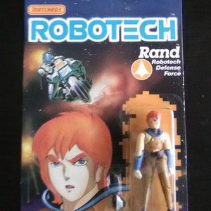 Puede incluir: Una figura de acción vintage de Robotech de Rand, miembro de la Fuerza de Defensa de Robotech. La figura está empaquetada en una caja de cartón azul y naranja con una imagen de Rand y un avión de combate Robotech. La caja también incluye el texto "Robotech" y "Rand".