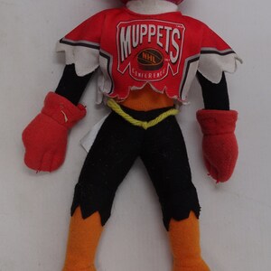 Può includere: Un giocattolo di peluche del personaggio Animal dei Muppets che indossa una maglia da hockey rossa e bianca con le parole "Muppets" e "NHL Conference". Il giocattolo ha i piedi arancioni e pantaloni neri.