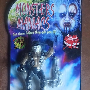 Puede incluir: Una figura de acción de Monsters Maniacs, empaquetada en una tarjeta azul y morada. La figura es un monstruo con sombrero negro, chaleco marrón y pantalones negros. La tarjeta presenta el texto "Get them before they get you!!!" y "Collect Them All!!".