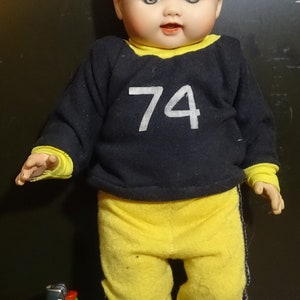 May include: Vintage baby doll in a navy and yellow outfit. The doll's top has the number 74 in white. It is wearing yellow trousers and shoes. A green lighter is next to the doll.