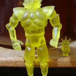 Puede incluir: Una figura de acción de plástico amarillo translúcido de un robot humanoide con una cabeza puntiaguda y un accesorio de cabeza puntiaguda separado. La figura tiene brazos y piernas articulados.