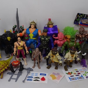 Op de afbeelding: Een verzameling vintage action figuren, waaronder een blauwe dinosaurus, een groene schildpad, een paars monster, een geel en oranje figuur en een vrouw in een rood, wit en blauw kostuum. De figuren zijn gerangschikt op een wit oppervlak met verschillende stickerbladen.