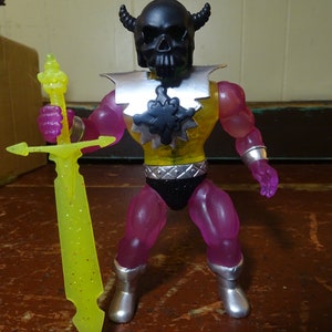 Puede incluir: Una figura de acción morada con un casco de calavera negro y una placa de pecho amarilla y plateada. La figura sostiene una espada amarilla con purpurina.