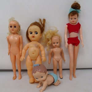 Puede incluir: Cinco muñecas de plástico vintage, incluyendo una muñeca bebé gateando a cuatro patas, una muñeca con cabello rubio y un vestido azul, una muñeca con cabello rubio y un vestido rojo, una muñeca con cabello castaño y un vestido rojo, y una muñeca con cabello rubio y un vestido azul.