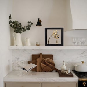 Op de afbeelding: Een gouden metalen olijftak-beeldhouwwerk in een zwart frame op een wit marmeren plank. Het beeldhouwwerk is omringd door andere woonaccessoires, waaronder een witte kan, een houten snijplank en een witte keramische pot.