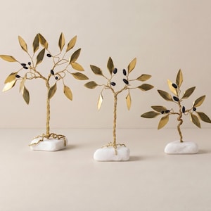 Peut inclure: Trois sculptures décoratives en forme d'arbres en métal. Chaque arbre présente des feuilles dorées et un tronc doré torsadé, avec de petits accents noirs. Les arbres sont montés sur des socles en pierre blancs et rectangulaires.