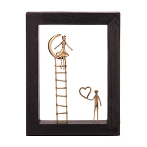 Op de afbeelding: Een metalen sculptuur van een stel in een zwart frame. De vrouw zit op een halve maan op een ladder, terwijl de man een hartvormige ballon vasthoudt. Het beeld is gemaakt van goudkleurig metaal.
