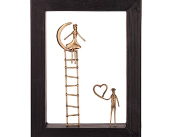 May include: A metal sculpture of a couple in a black frame. The woman is sitting on a crescent moon on a ladder, while the man is holding a heart-shaped balloon. The sculpture is made of gold-colored metal.