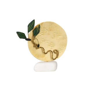 May include: A decorative sculpture featuring a textured gold-colored disc with a green and gold leaf design and a twisted gold-colored wire. The sculpture is balanced on a white stone base. The leaves have a dark green and gold pattern.