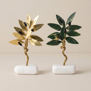 May include: Two decorative olive tree sculptures. One is gold-colored, the other is green. Both have black olive accents and are mounted on white rectangular bases. The stems are twisted wire.