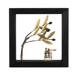 Op de afbeelding: Een zwarte ingelijste wandsculptuur met een gouden metalen olijftak met zwarte olijven en een stel dat op een bankje onder de tak zit.