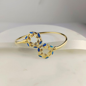 Op de afbeelding: Een gouden armband met een dubbele bladeren ontwerp. De bladeren zijn omrand met goud en gevuld met blauw email.