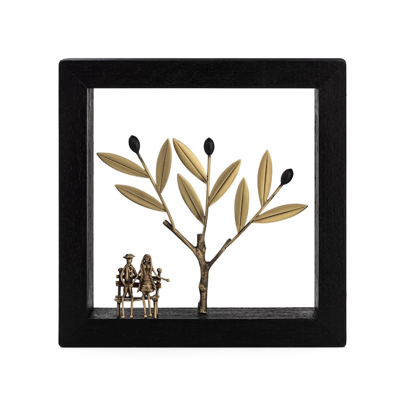 Op de afbeelding: Een zwarte ingelijste wanddecoratie met een gouden metalen olijfboom met zwarte olijven en een stel dat op een bankje onder de boom zit.