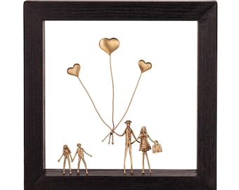 May include: A square black wooden frame containing a bronze-colored metal sculpture of a family of four. Two adults and two children are depicted standing, with the adults holding three heart-shaped balloons on thin wires above their heads.