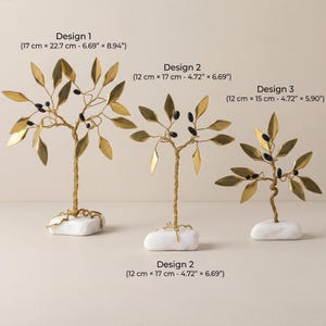 May include: Three decorative golden metal tree sculptures with leaf and berry details, each mounted on a white stone base. Design 1 measures 6.69" x 8.94", Design 2 measures 4.72" x 6.69", and Design 3 measures 4.72" x 5.90".